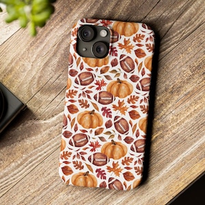 May include: A white iPhone case featuring a repeating pattern of orange pumpkins, brown footballs, and autumn leaves. The case has a glossy finish and is designed to fit a modern smartphone. The design evokes a fall or harvest theme.
