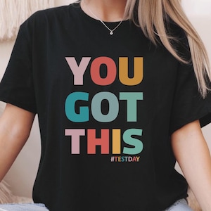 May include: Black t-shirt with the words "YOU GOT THIS" in large, colorful letters. The text is in pink, red, teal, yellow, and pink. Below the text is the hashtag "#TESTDAY".