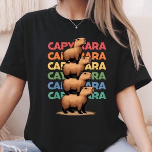 May include: Black t-shirt with a stack of five capybaras in a rainbow gradient pattern. The text "CAPYBARA" is repeated five times in a rainbow gradient pattern.
