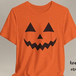 Może przedstawiać: Pomarańczowa koszulka z czarnym wzorem twarzy dyni. Wzór przedstawia trójkątne oczy, trójkątny nos i poszarpaną buzię. Słowa "heather orange" są napisane po prawej stronie.