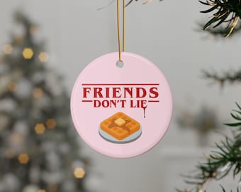 Puede incluir: Adorno navideño redondo y rosa con el texto rojo "FRIENDS DON'T LIE". Debajo, un gráfico de un gofre con mantequilla en un plato. El adorno cuelga de un hilo dorado.