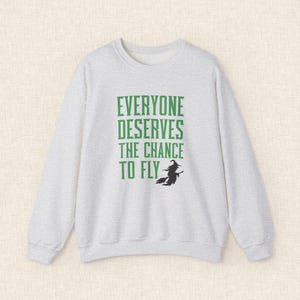 Op de afbeelding: Een lichtgrijze sweatshirt met de groene tekst "EVERYONE DESERVES THE CHANCE TO FLY". Een zwart silhouet van een heks op een bezem staat onder de tekst. De sweatshirt heeft een ronde hals en lange mouwen.