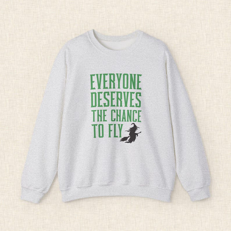 Op de afbeelding: Een lichtgrijze sweatshirt met de groene tekst "EVERYONE DESERVES THE CHANCE TO FLY". Een zwart silhouet van een heks op een bezem staat onder de tekst. De sweatshirt heeft een ronde hals en lange mouwen.