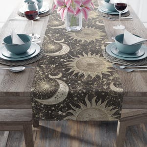 May include: A celestial-themed table runner featuring sun and moon designs in gold against a dark background. The runner is placed on a wooden table set with plates, bowls, wine glasses, and silverware, creating an elegant dining setting.