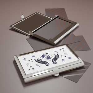 Pode incluir: Um porta-cartões de metal prateado com uma parte superior branca com um design celestial com mãos, estrelas e um cristal. O porta-cartões é retangular e tem uma tampa articulada. O design é em tons de roxo e preto.