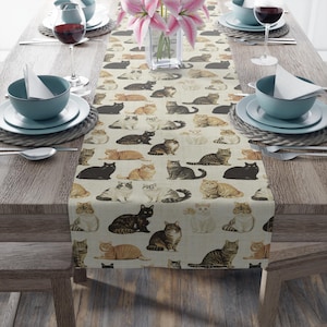 May include: A beige table runner featuring a repeating pattern of various cat breeds in different poses. The cats are depicted in shades of black, brown, gray, and orange. The runner is placed on a wooden table set for a meal.