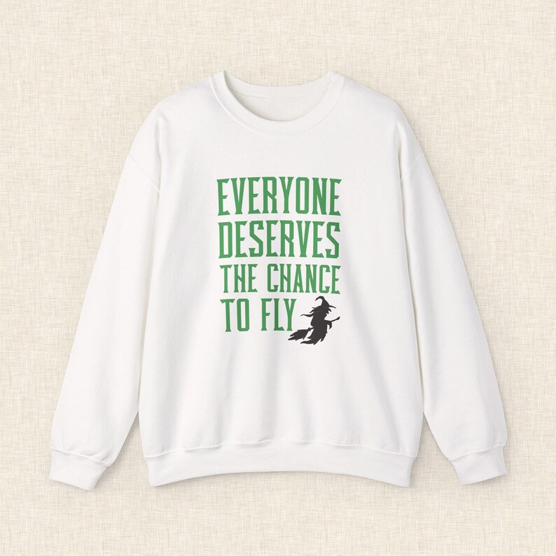 Op de afbeelding: Witte sweatshirt met de groene tekst "EVERYONE DESERVES THE CHANCE TO FLY" en een zwart heksen silhouet. De top met lange mouwen en ronde hals is een casual kledingstuk.