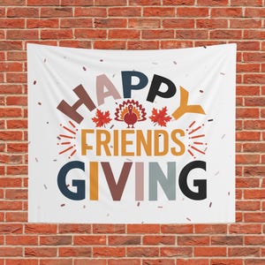May include: A white fabric wall hanging featuring the text HAPPY FRIENDS GIVING in large, multi-colored block letters. The design includes a central graphic of a turkey and two red maple leaves, surrounded by small scattered brown confetti shapes.