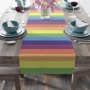 Puede incluir: Un camino de mesa a rayas de arcoíris con colores alternos de rojo, naranja, amarillo, verde, azul, añil y violeta. El camino de mesa está colocado sobre una mesa de madera con cubiertos a cada lado.
