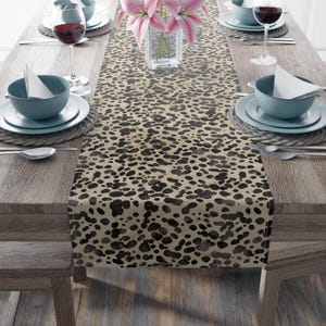 May include: A table runner with a beige background and a black and brown spotted pattern. The runner is placed on a wooden table set for a meal, with plates, bowls, silverware, and wine glasses.