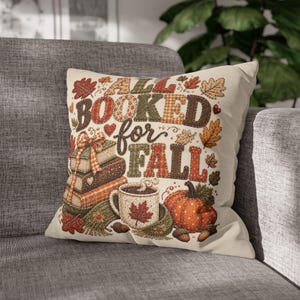 May include: A square throw pillow with a cream background featuring the text ALL BOOKED for FALL in multicoloured, patterned letters. The design includes a stack of books with a plaid ribbon, a mug with a maple leaf, a pumpkin, and acorns.