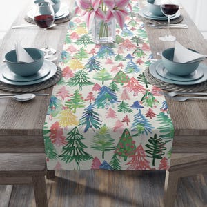 May include: A white table runner with a colorful watercolor pattern of Christmas trees. The trees are painted in shades of green, blue, red, pink, and yellow. The runner is perfect for adding a festive touch to your holiday table.