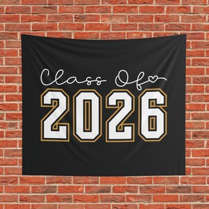 May include: A black rectangular fabric banner featuring the white cursive text Class Of followed by a small heart icon. Below the text, the large numbers 2026 are printed in a white collegiate font with a gold outline, centred on the black background.