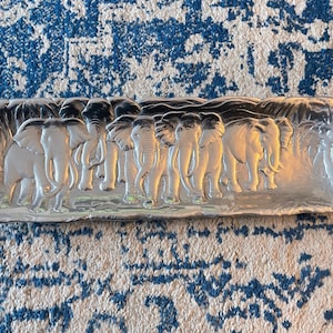 May include: A rectangular, silver-colored tray with a raised relief of a herd of elephants. The elephants are depicted in various sizes and poses, with detailed trunks and ears. The tray has a slightly curved edge.