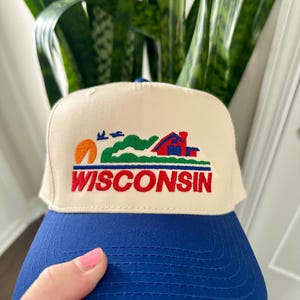May include: A cream-colored trucker hat with a blue brim. The hat features an embroidered design of a sunset, rolling hills, a red house, and the word "WISCONSIN" in red letters.