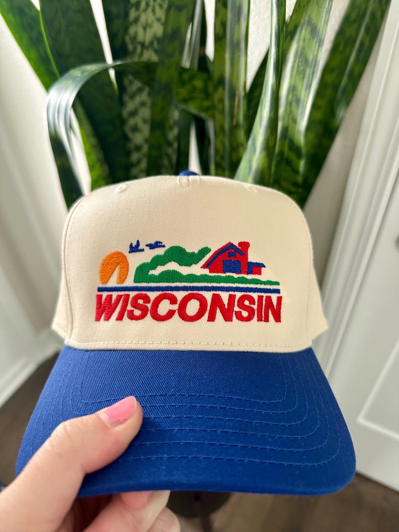May include: A cream-colored trucker hat with a blue brim. The hat features an embroidered design of a sunset, rolling hills, a red house, and the word "WISCONSIN" in red letters.