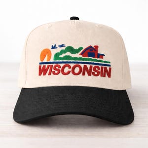 May include: Cream-colored baseball cap with a black brim. The cap features an embroidered design of a red house, green hills, a setting sun, and the word "WISCONSIN" in red and blue.