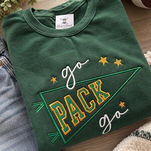 May include: A dark green crewneck sweatshirt featuring embroidered text that reads "go PACK go" in white and gold lettering. The design includes a green pennant flag outline and three small gold stars. A white brand label reads "Loop & Stitch".