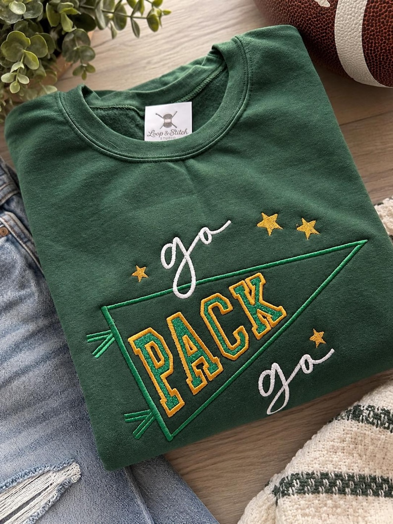 May include: A dark green crewneck sweatshirt featuring embroidered text that reads "go PACK go" in white and gold lettering. The design includes a green pennant flag outline and three small gold stars. A white brand label reads "Loop & Stitch".