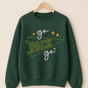 May include: A dark green crewneck sweatshirt featuring an embroidered design with the text "go PACK go" in white and yellow. The design includes a green pennant flag outline and three yellow embroidered stars on a long-sleeve pullover garment.