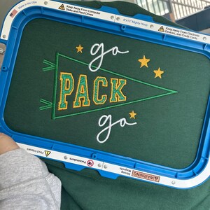 May include: A dark green fabric held in a blue 8x13 inch Mighty Hoop embroidery frame. The fabric features an embroidered green pennant with yellow glittery text reading PACK, surrounded by white cursive text reading go and yellow stars.