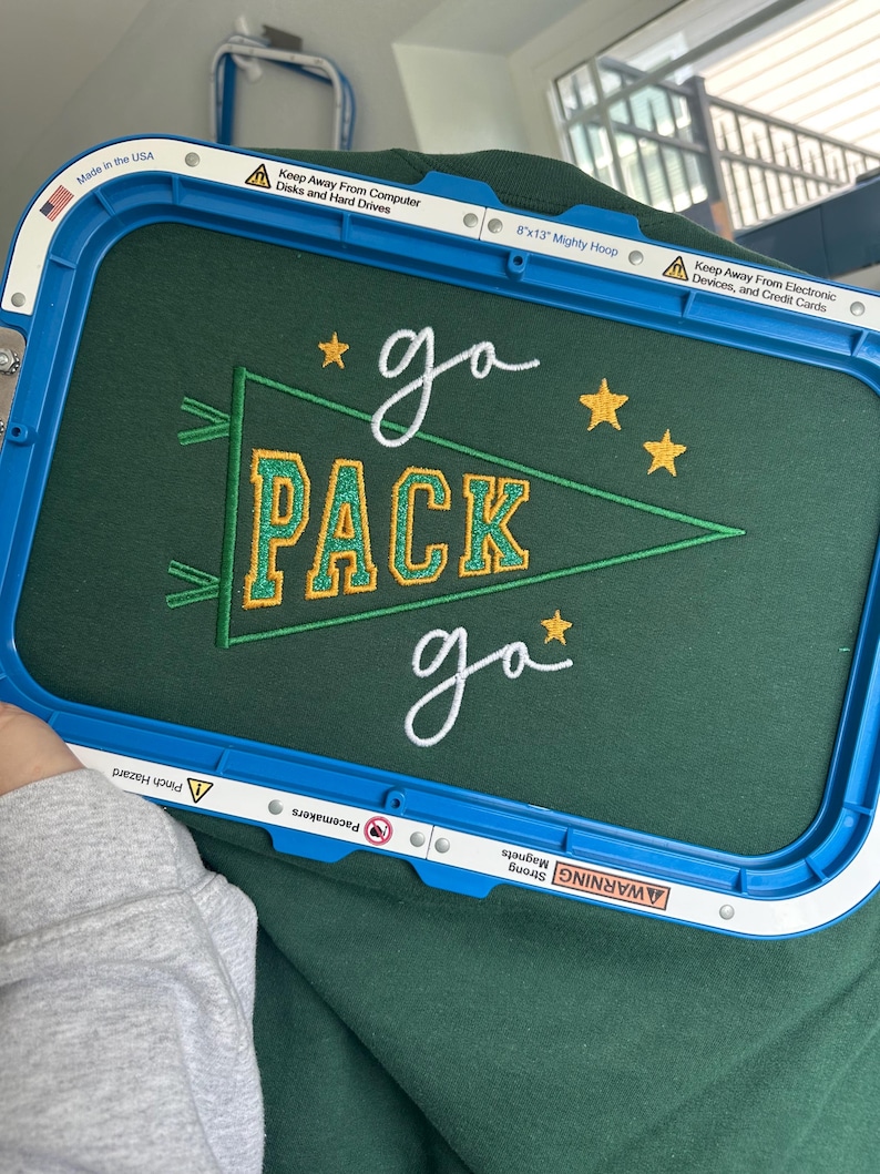 May include: A dark green fabric held in a blue 8x13 inch Mighty Hoop embroidery frame. The fabric features an embroidered green pennant with yellow glittery text reading PACK, surrounded by white cursive text reading go and yellow stars.