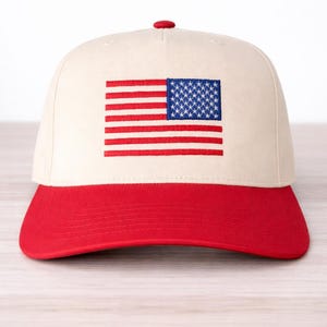 May include: A baseball cap with a cream-colored crown and a red brim. The front of the cap features an embroidered American flag. The flag has red and white stripes and a blue square with white stars.
