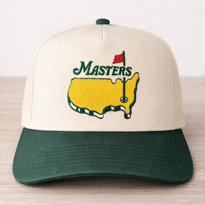 May include: A cream-colored baseball cap with a green brim and top button. The cap features the word "Masters" in green script above a yellow embroidered outline of the state of Georgia, with a red flag and golf pin.