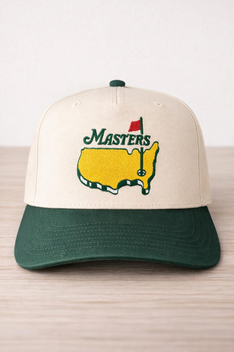 May include: A cream-colored baseball cap with a green brim and top button. The cap features the word "Masters" in green script above a yellow embroidered outline of the state of Georgia, with a red flag and golf pin.