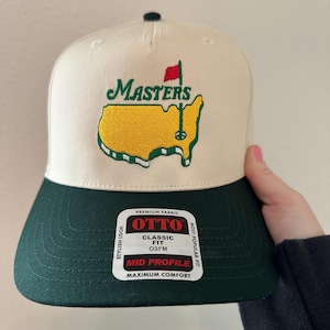 May include: A cream-colored baseball cap with a green brim and a green and yellow embroidered logo. The logo features the word "MASTERS" above a yellow outline of a golf course with a red flag. The cap has a "OTTO" tag.