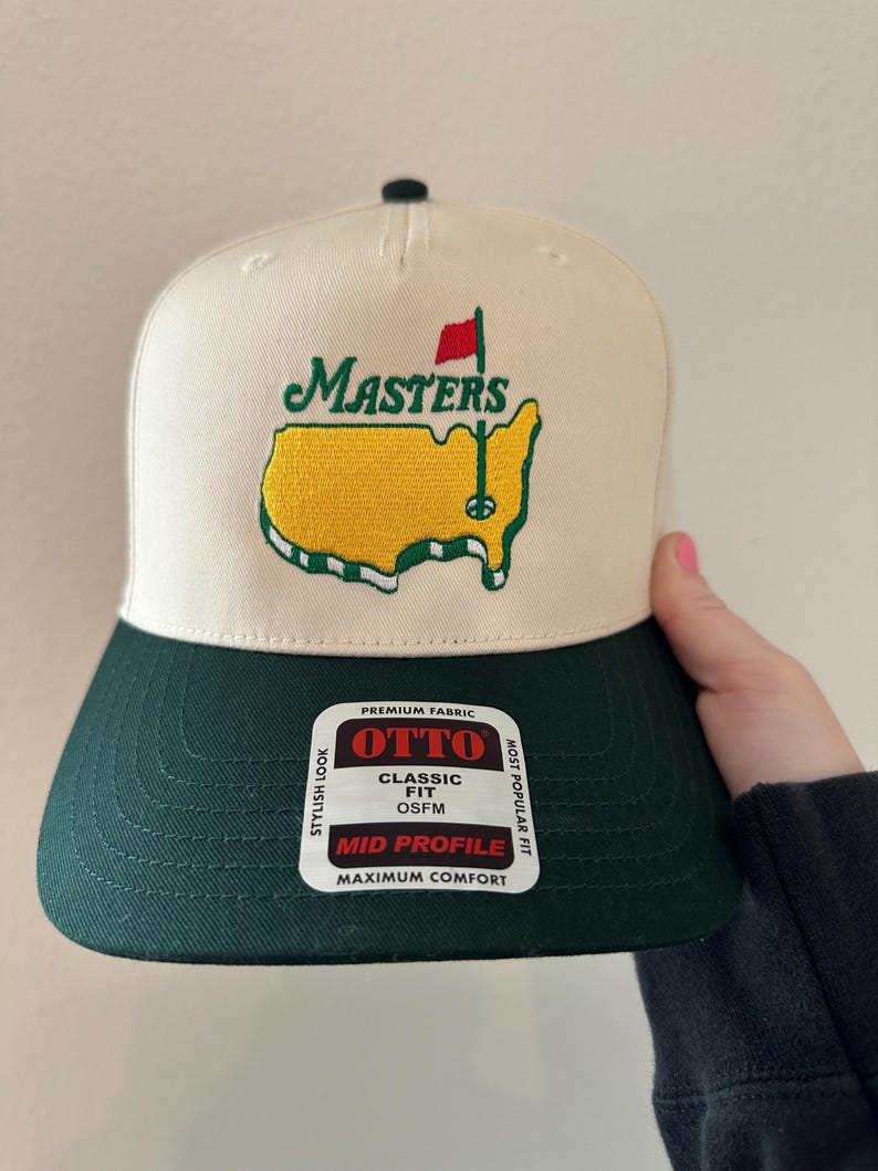 May include: A cream-colored baseball cap with a green brim and a green and yellow embroidered logo. The logo features the word "MASTERS" above a yellow outline of a golf course with a red flag. The cap has a "OTTO" tag.