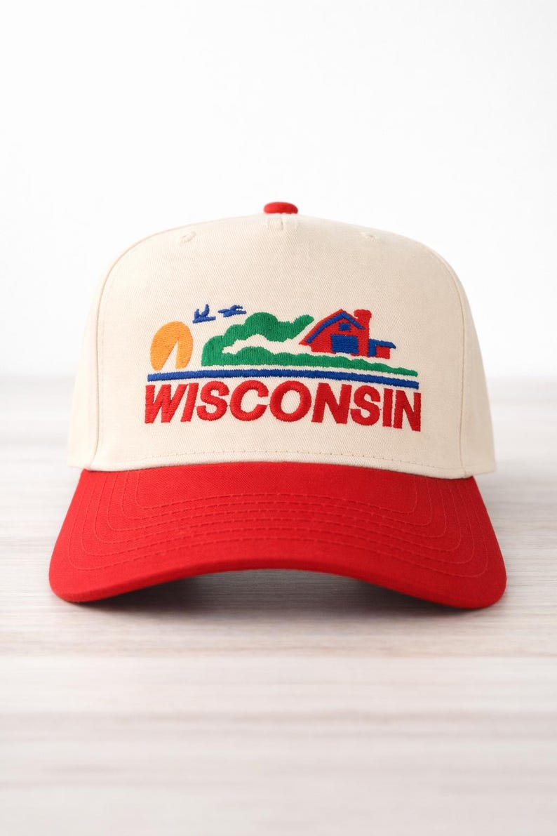 May include: A cream-colored baseball cap with a red brim. The cap features an embroidered design with a sunset, birds, and a house, with the word "WISCONSIN" in red block letters.