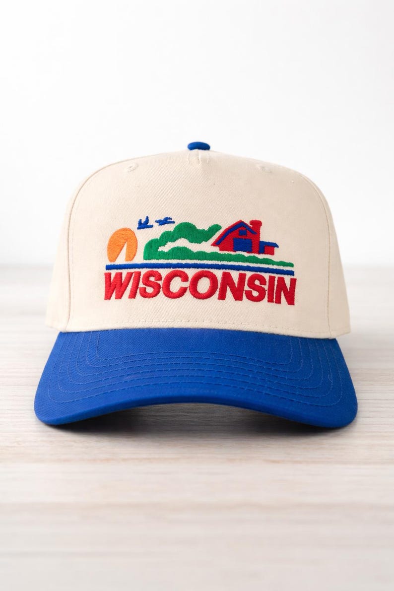 May include: A cream-colored baseball cap with a blue brim. The cap features an embroidered design of a sunset, a house, and the word "WISCONSIN" in red. The cap is a classic, structured style.