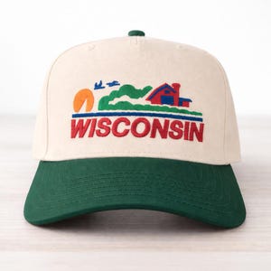 May include: Cream-colored baseball cap with a green brim. The cap features an embroidered design of a red house, green hills, an orange sun, and the word "WISCONSIN" in red letters.