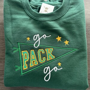 May include: A forest green crewneck sweatshirt featuring an embroidered design of a pennant flag with the text "PACK" in yellow and green. The words "go" are embroidered in white above and below the pennant, accented by small yellow stars.