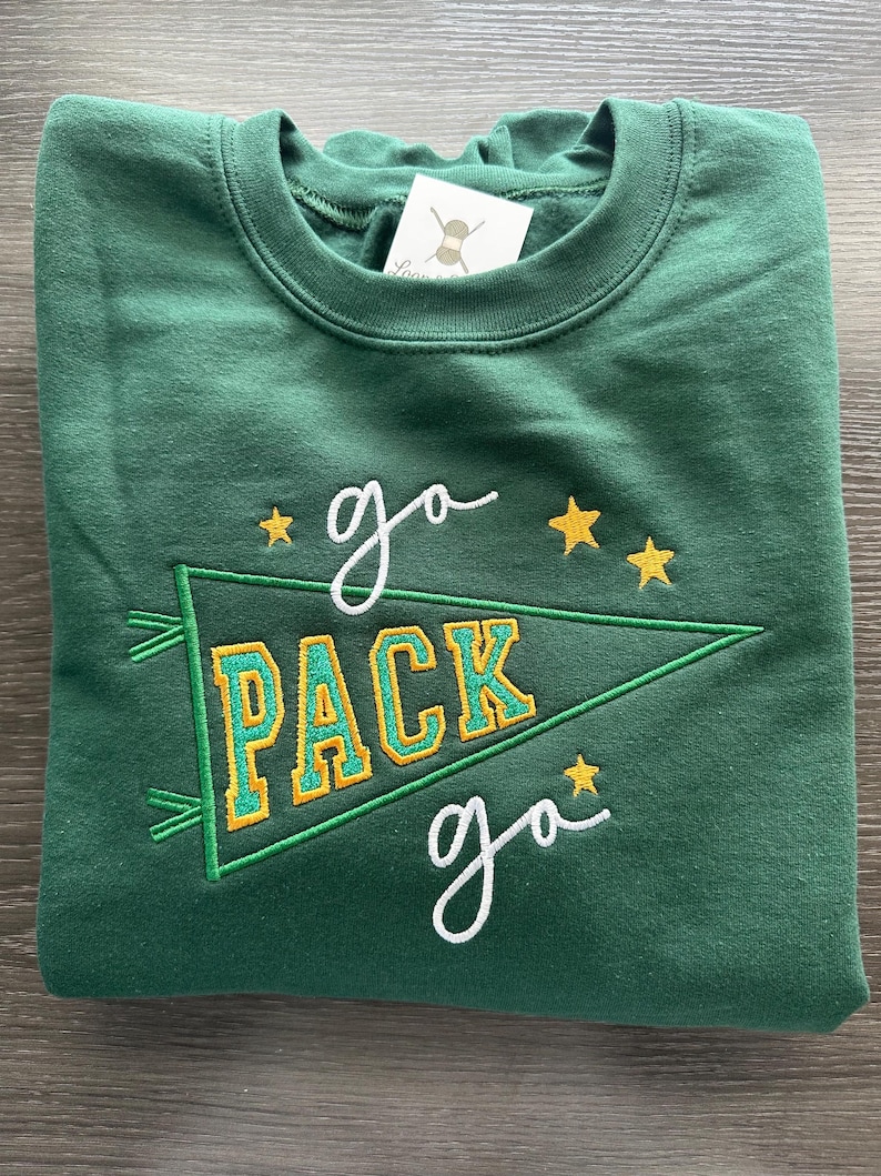 May include: A forest green crewneck sweatshirt featuring an embroidered design of a pennant flag with the text "PACK" in yellow and green. The words "go" are embroidered in white above and below the pennant, accented by small yellow stars.