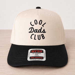 May include: A beige and black baseball cap with the text "COOL Dads CLUB" embroidered in black. The cap has a black brim and a small black button on top. The cap is made of a premium fabric and is a classic fit.