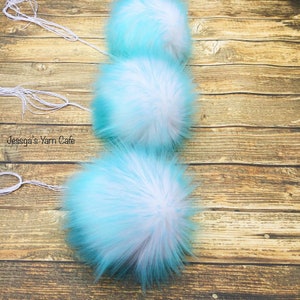 May include: Three fluffy pom poms in shades of blue and white, with white string attached to each. The pom poms are arranged vertically on a wooden surface.