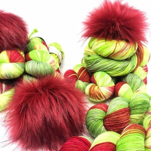 May include: Two skeins of green, red, and yellow yarn with red faux fur pom poms. The yarn is a variegated colorway.