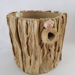 Puede incluir: Una maceta de cerámica con forma de tronco de árbol con un agujero en el lateral. La maceta está decorada con pequeños insectos de cerámica.