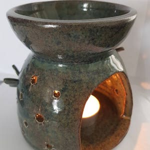 Puede incluir: Un quemador de aceite de cerámica verde con un cuenco redondo en la parte superior y un recorte para una vela de té. El quemador tiene un esmalte moteado y un diseño de pequeños agujeros.