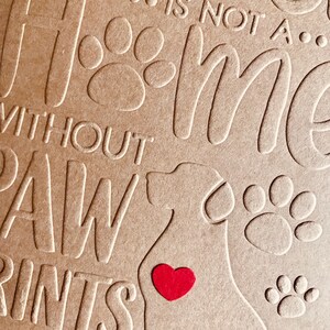 Puede incluir: Una tarjeta marr&oacute;n con el texto "Home is not a... without paw prints" y una silueta de un perro con un coraz&oacute;n rojo en el pecho. La tarjeta est&aacute; decorada con huellas de patas.