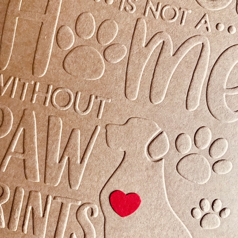 Puede incluir: Una tarjeta marr&oacute;n con el texto "Home is not a... without paw prints" y una silueta de un perro con un coraz&oacute;n rojo en el pecho. La tarjeta est&aacute; decorada con huellas de patas.
