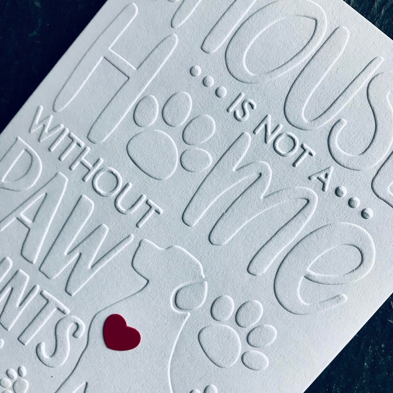 Op de afbeelding: Witte kaart met reli&euml;ftekst die "Home is not a house without paw prints" luidt. De kaart heeft een rood hart en potafdrukken.