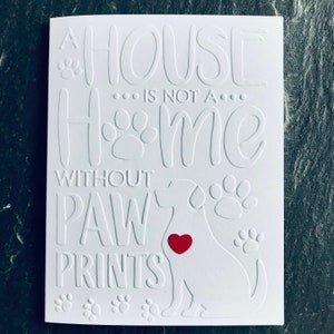 Op de afbeelding: Witte kaart met reli&euml;ftekst die luidt "Een huis is geen thuis zonder poot afdrukken". Een wit silhouet van een hond met een rood hart bevindt zich in het midden van de kaart.
