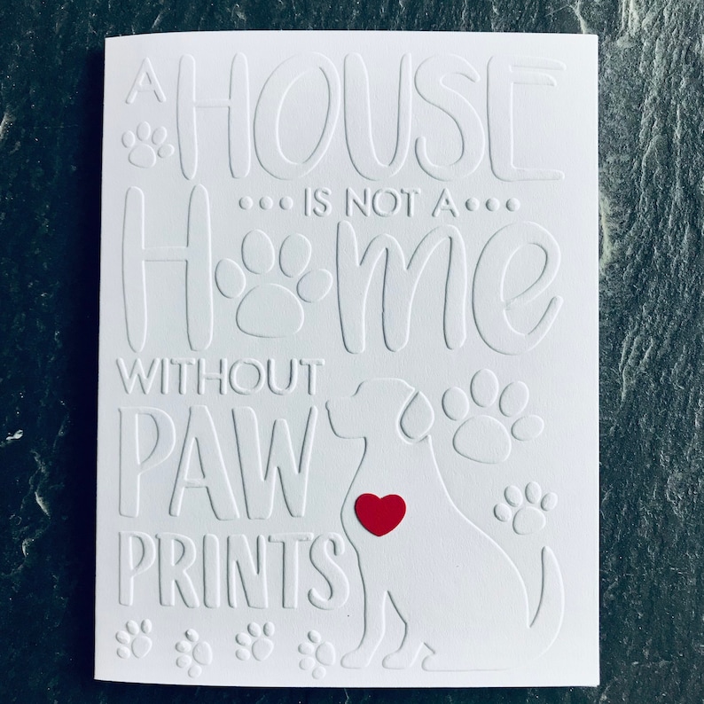 Op de afbeelding: Witte kaart met reli&euml;ftekst die luidt "Een huis is geen thuis zonder poot afdrukken". Een wit silhouet van een hond met een rood hart bevindt zich in het midden van de kaart.