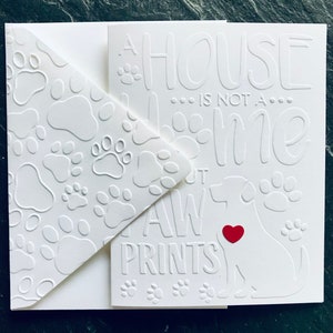 Op de afbeelding: Een witte kaart met een pootprintpatroon aan de linkerkant. De rechterkant heeft de tekst "Een huis is geen thuis zonder pootprinten" met een wit silhouet van een hond met een rood hart in het midden.
