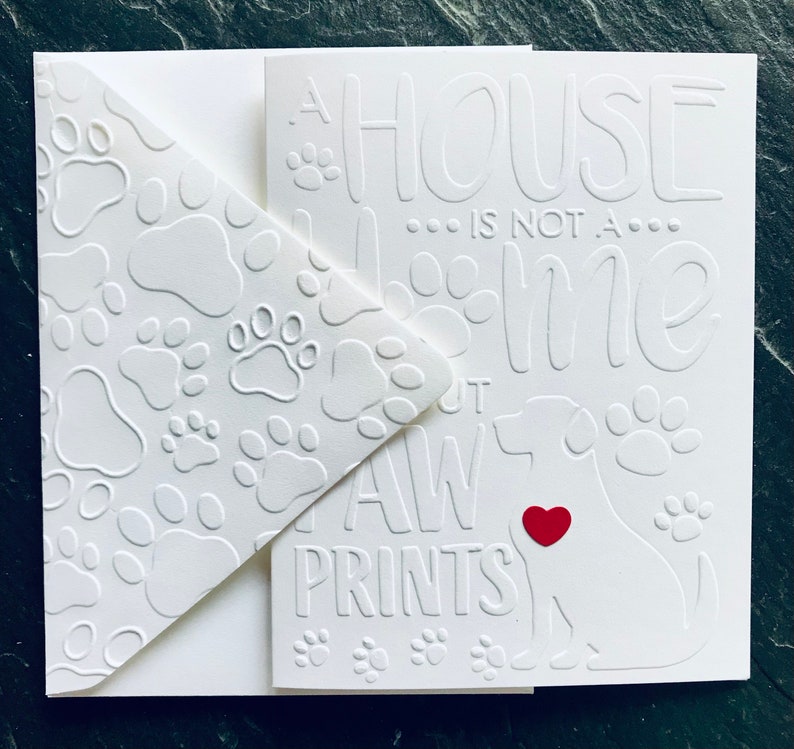 Op de afbeelding: Een witte kaart met een pootprintpatroon aan de linkerkant. De rechterkant heeft de tekst "Een huis is geen thuis zonder pootprinten" met een wit silhouet van een hond met een rood hart in het midden.