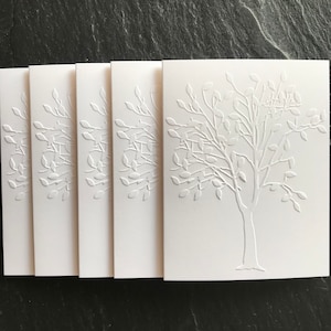 May include: A stack of five white cards with an embossed tree design. The tree features a trunk and branches with leaves. The cards are arranged vertically, with the tree design centered on each card. The cards are made of a thick, textured paper.