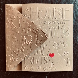 Puede incluir: Una tarjeta marr&oacute;n con un patr&oacute;n de huellas de patas en el lado izquierdo. El lado derecho tiene el texto "A house... is not a... home... Paw Prints" con una silueta de un perro con un coraz&oacute;n rojo en el pecho.